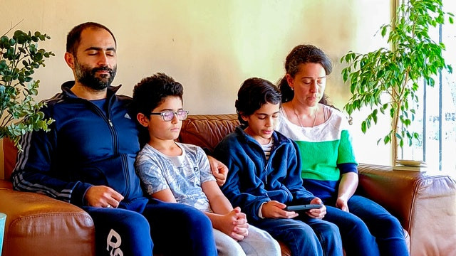 a family of four is sitting on the couch praying, the two young boys in the corner and the mother and father on the two corners of the couch