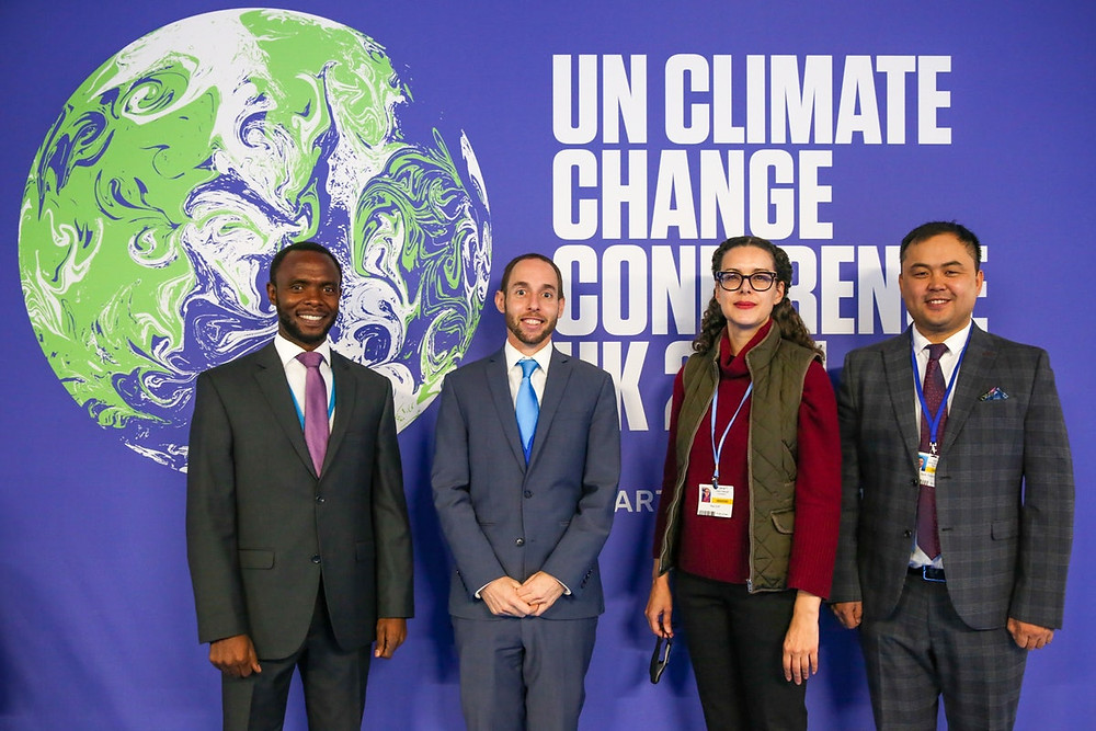 three people, two men and one women are shown to be sitting at a rectangular table in front of a screen that says "Strengthening Global Climate Governance"