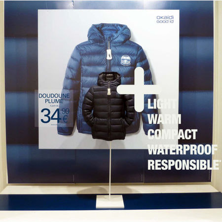Scénographie de vitrine technique hivernale mettant en avant une doudoune en plume Okaïdi sur fond bleu.