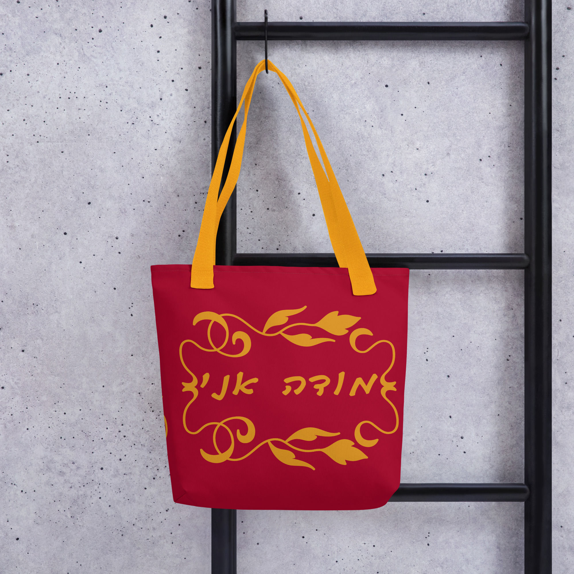 A carmine tote bag with yellow handles on hanger hooked onto a ladder-like metal structure with the modeh ani design