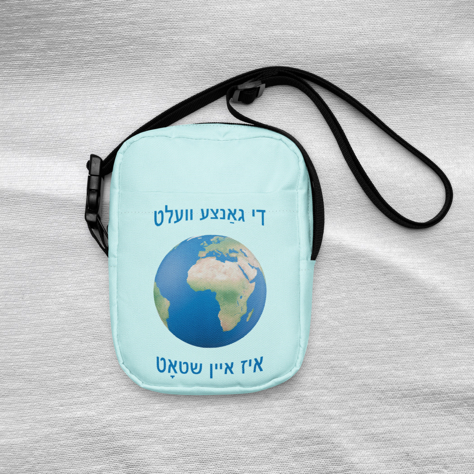 Light blue crossbody bag the front laying. Design the Yiddish phrase in blue Hebrew letters 2 rows + the globe in between