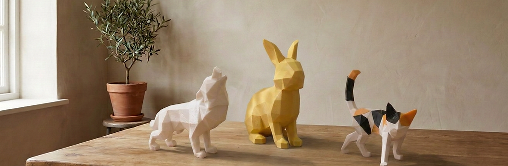 Three low-poly animal figurines—a white wolf, yellow rabbit, and calico cat—displayed on a warm oak worktable in a softly lit creative studio.