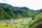 Campsite with tents and vehicles in a green valley surrounded by hills. People are scattered around. A small stream runs nearby.