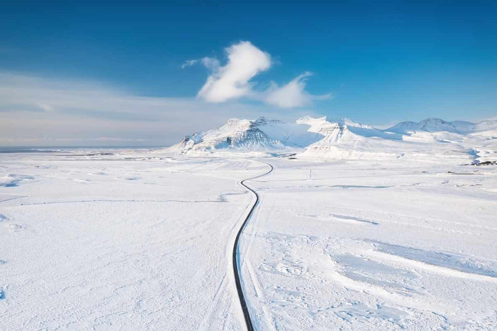 A winding road cuts through a vast snowy landscape, leading to distant mountains under a bright blue sky with a few clouds.