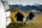 Two people in winter wear sit on chairs by a campervan, overlooking snowy mountains. Cars parked nearby under a cloudy sky. Relaxed vibe.