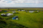 Green campsite scene with tents, caravans, and cars under cloudy skies. People are gathered near a blue tent, suggesting a relaxed atmosphere.