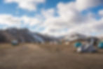 Tents and cars in a rocky campsite with mountains and snow patches under a cloudy blue sky, in landmannalaugar.