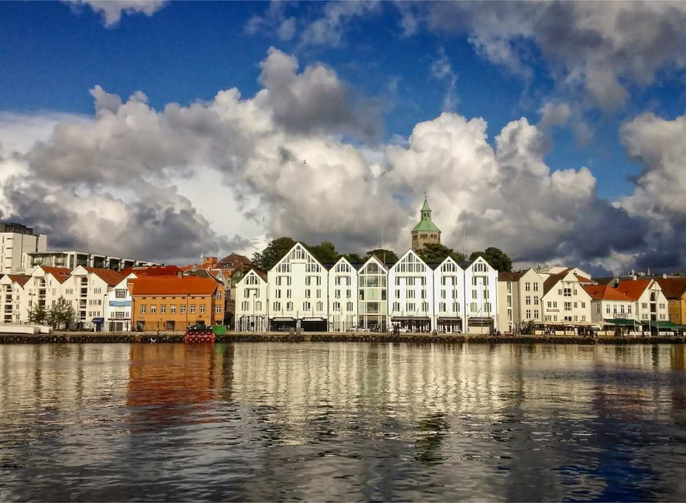 White and orange buildings line a waterfront of Stavanger under a sky filled with dramatic clouds. A tower with a green top is visible.