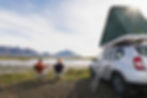Two people sit on chairs by a lake, toasting mugs. A car with a rooftop tent is nearby. Mountains in the background on a sunny day.