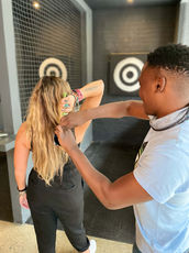 Man in a black t-shirt aiming an axe toward the camera during an indoor axe throwing session.