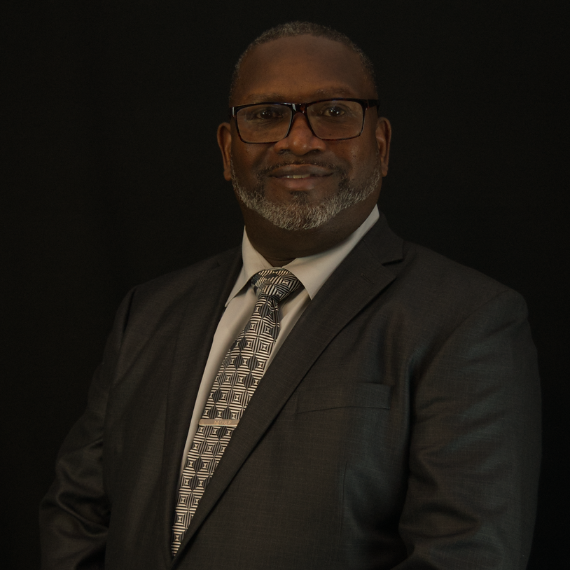 Black man in a grey suit and brown tie against a dark background