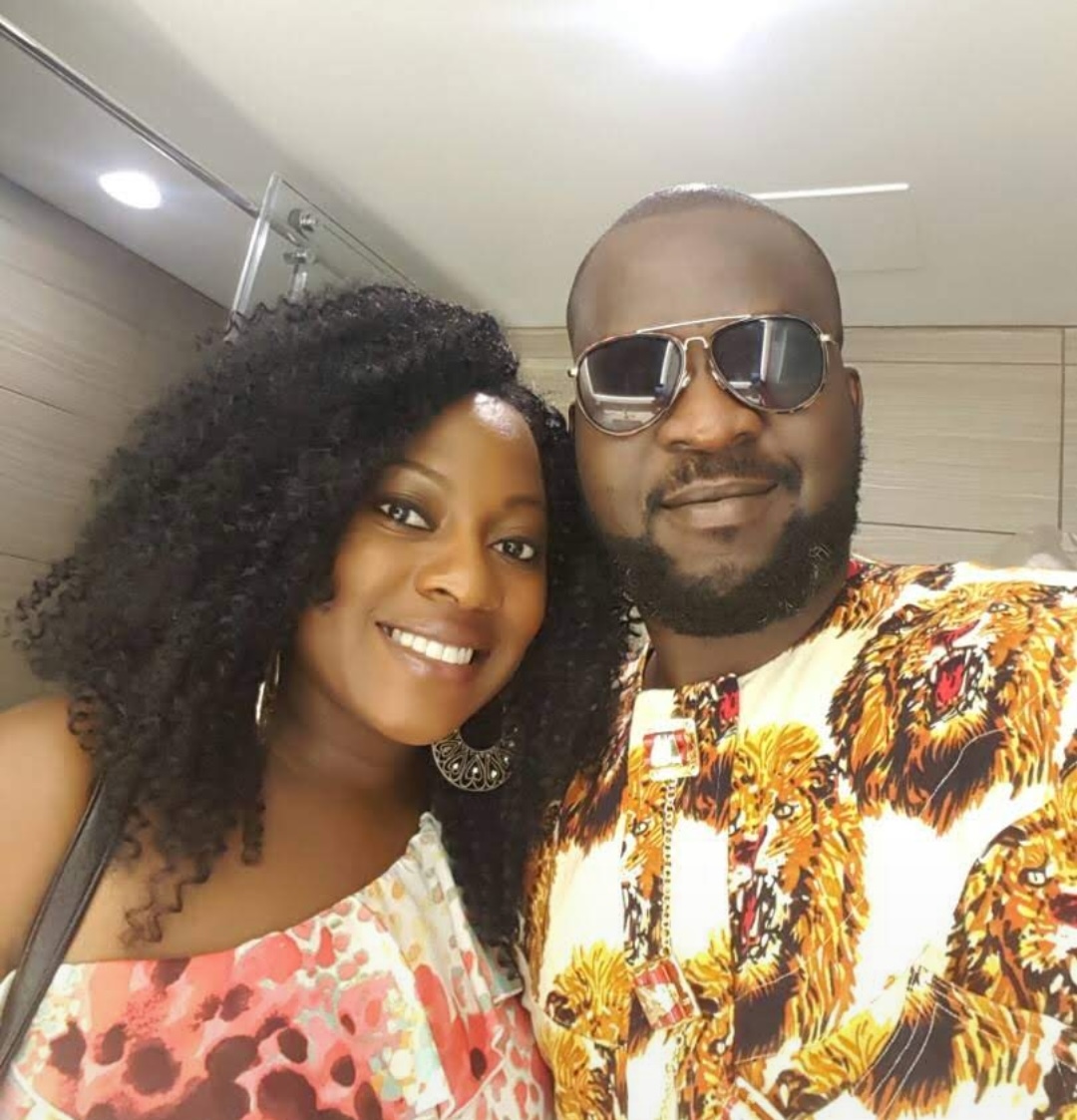 Smiling couple taking a selfie indoors; woman with curly hair in a floral dress, man in sunglasses and a lion-print shirt.