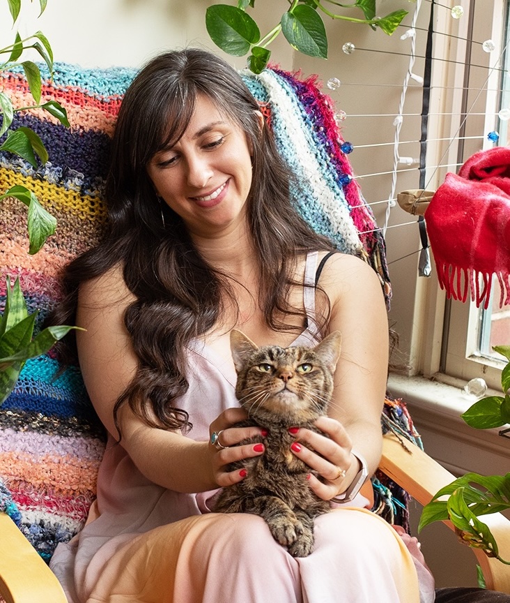 Smiling woman in a pink dress holds a tabby cat on her lap by a window, wrapped in a colorful crocheted blanket with houseplants.