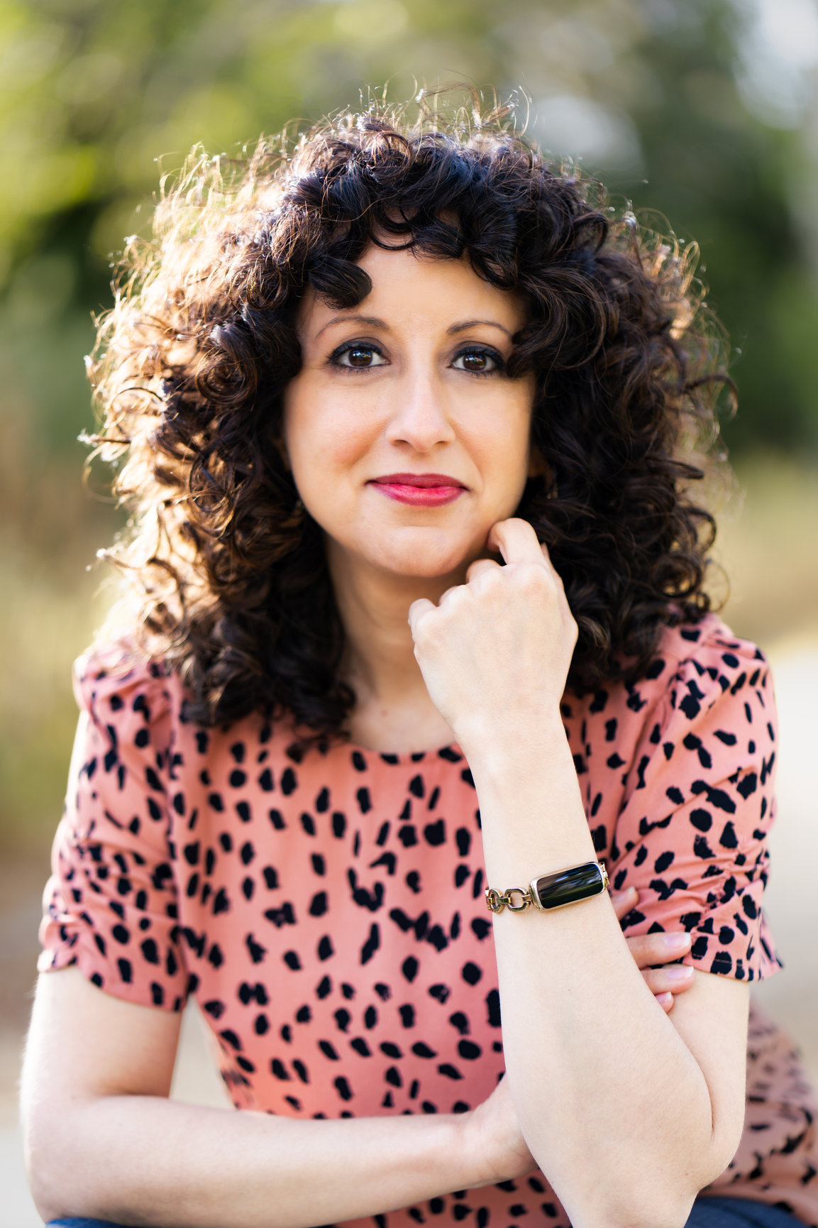 Portrait of a woman Olivia Abousaid with curly dark hair in a pink patterned top, resting her chin on her hand outdoors.