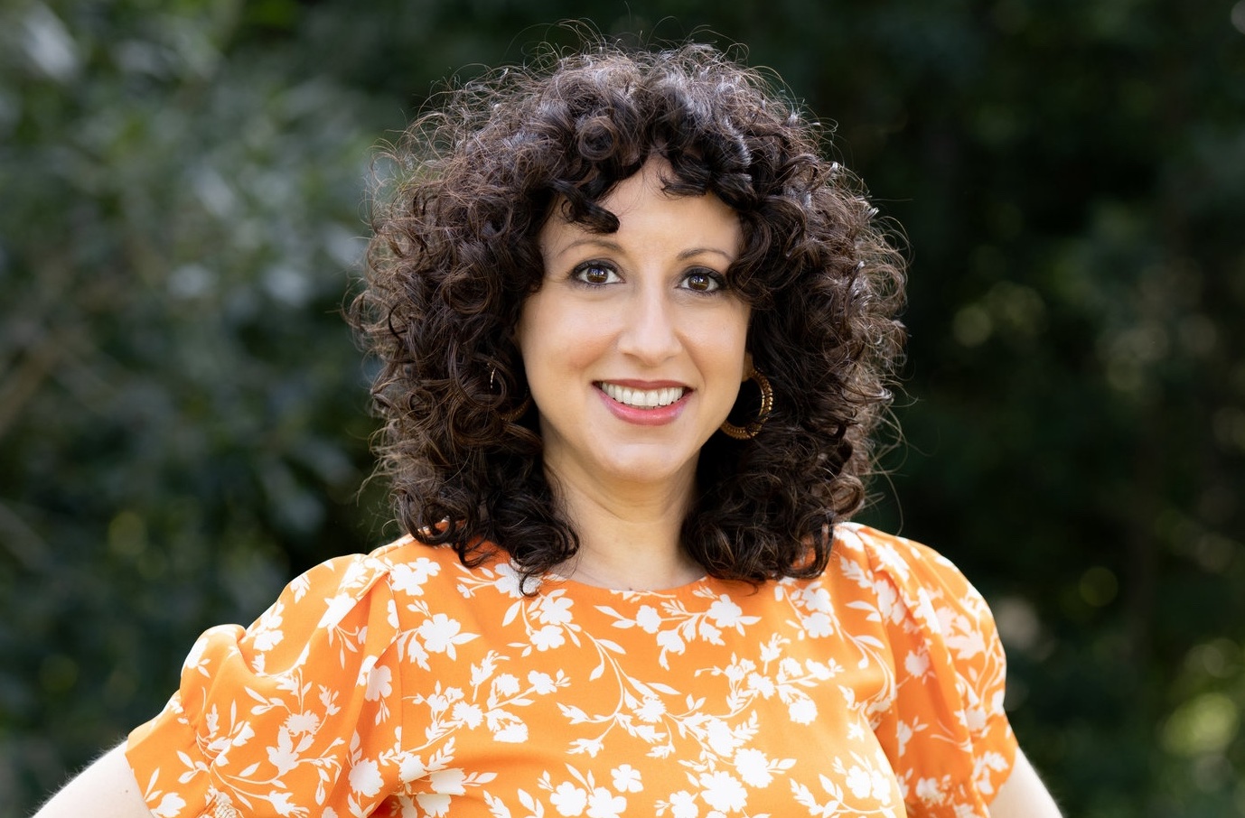 Smiling woman Olivia Abousaid with curly dark hair in an orange floral dress stands outdoors against a blurred green background.