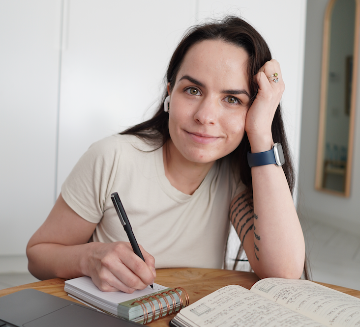 Woman in a beige shirt smiles while writing in a notebook at a table, with a laptop and open planner nearby.