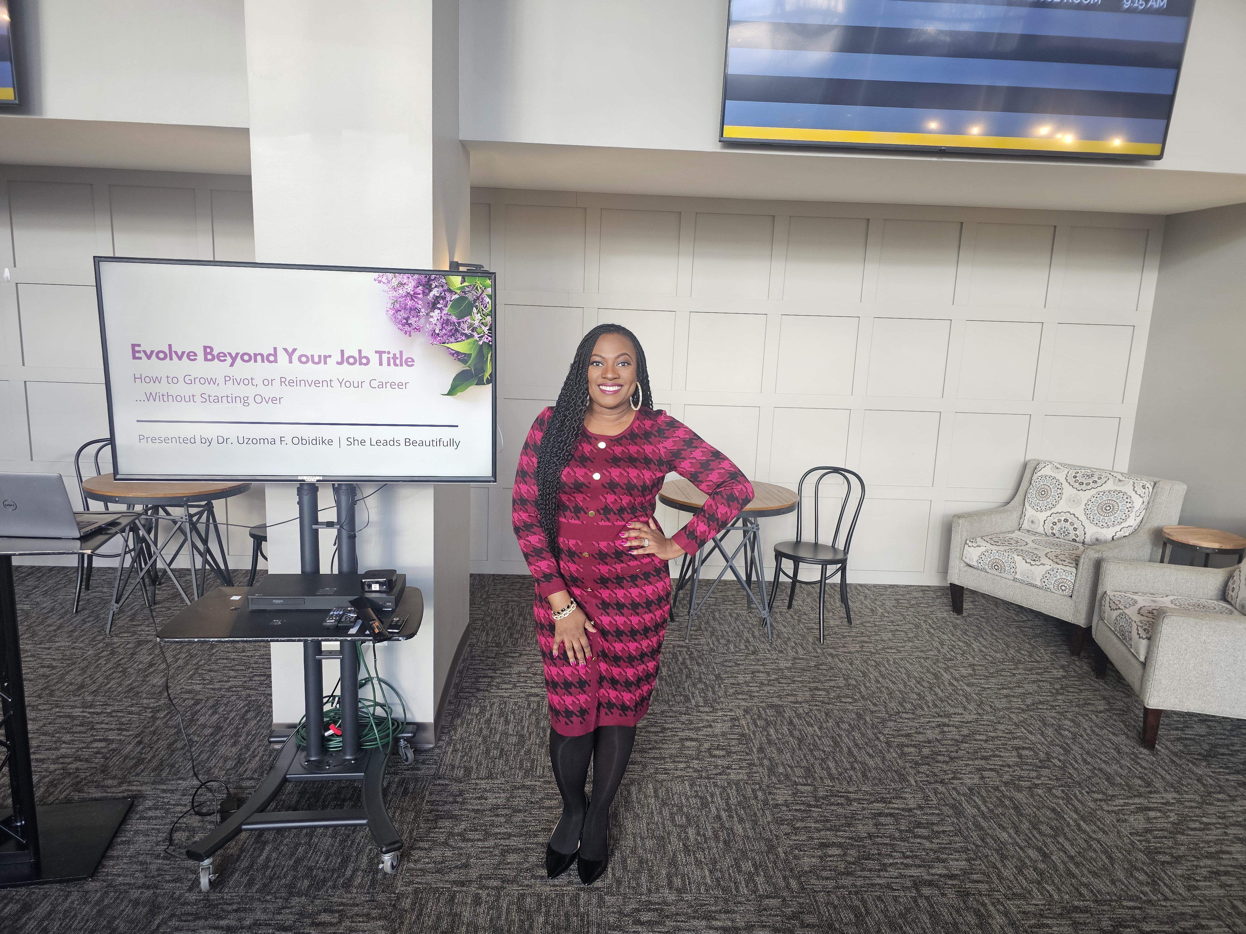 Smiling woman Uzoma Obidike in a magenta dress stands beside a screen reading Evolve Beyond Your Job Title in a modern lounge-like room.