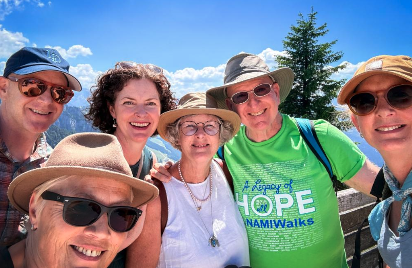 Six smiling people in hats and sunglasses pose for a selfie at a mountain overlook under a bright blue sky.