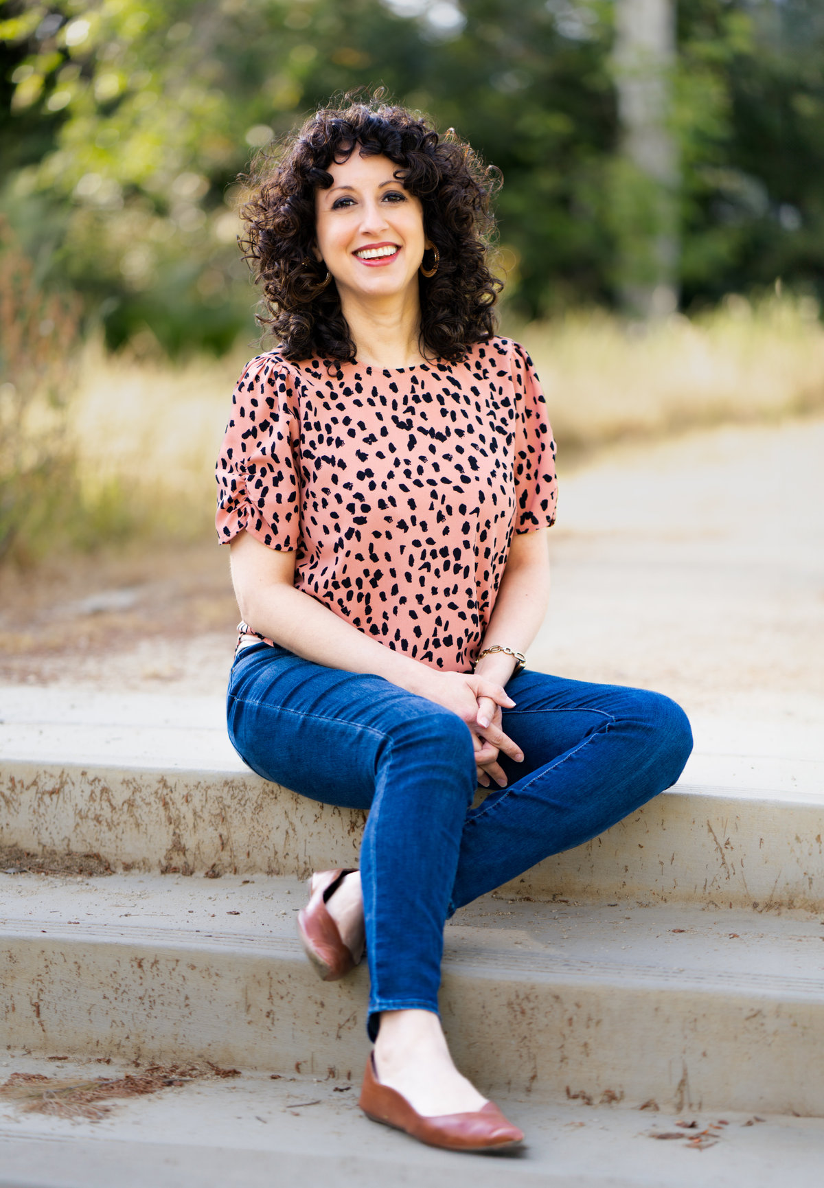 Smiling woman Olivia Abousaid with curly hair sits on outdoor steps in a pink leopard-print top and blue jeans, with a blurred green background.