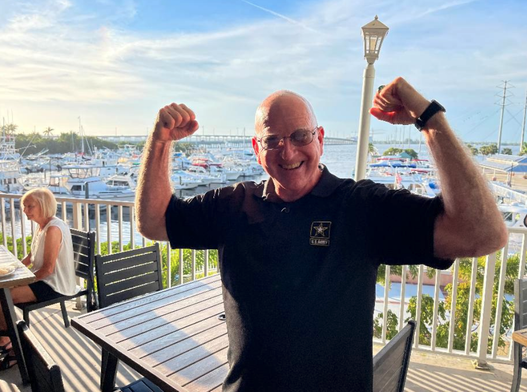 Smiling man in black polo flexes at a sunny marina deck, with boats, a bridge, and seated woman in the background.