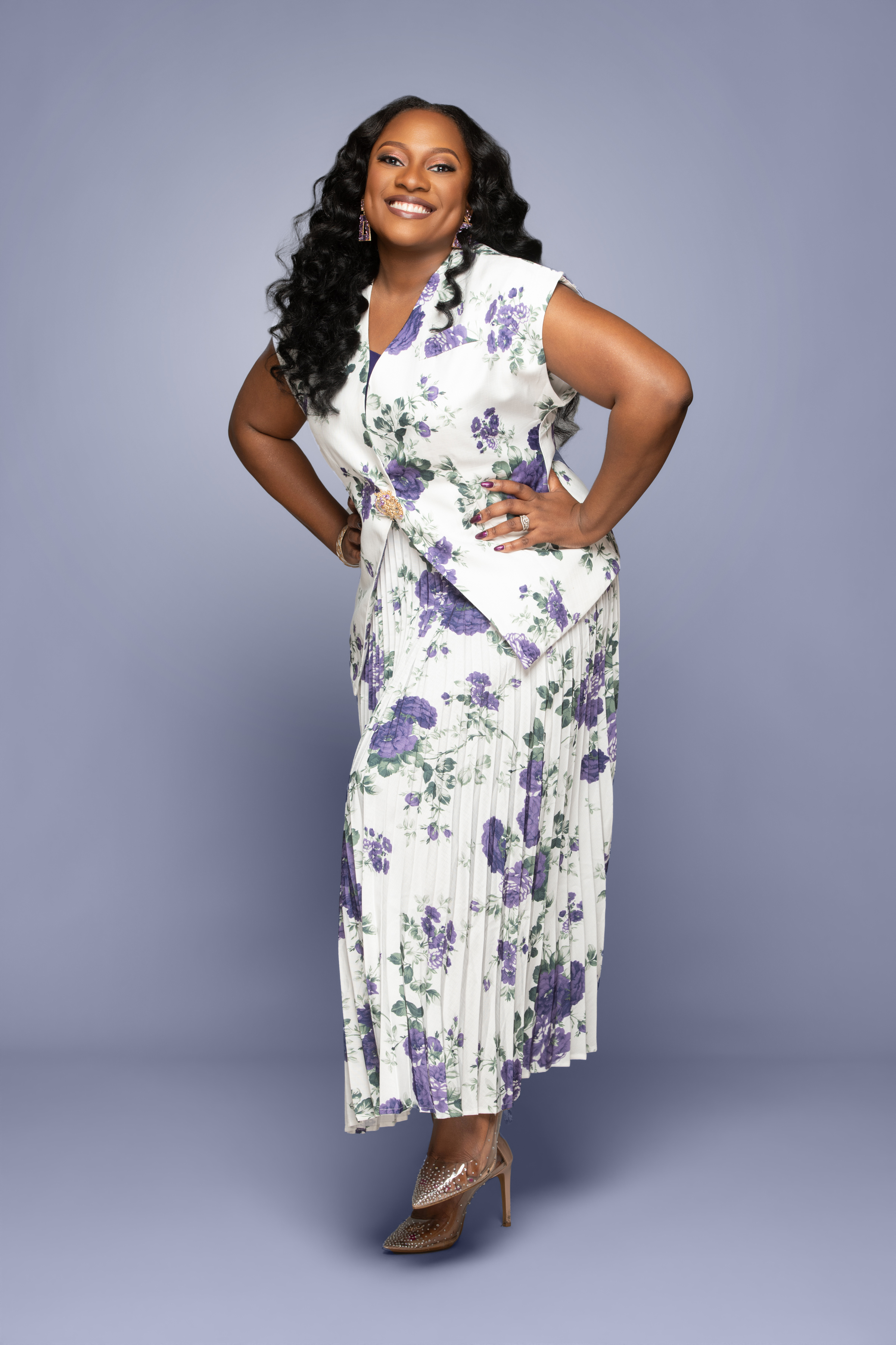 Smiling woman Uzoma Obidike poses in a white-and-purple floral dress and heels against a plain gray studio backdrop.