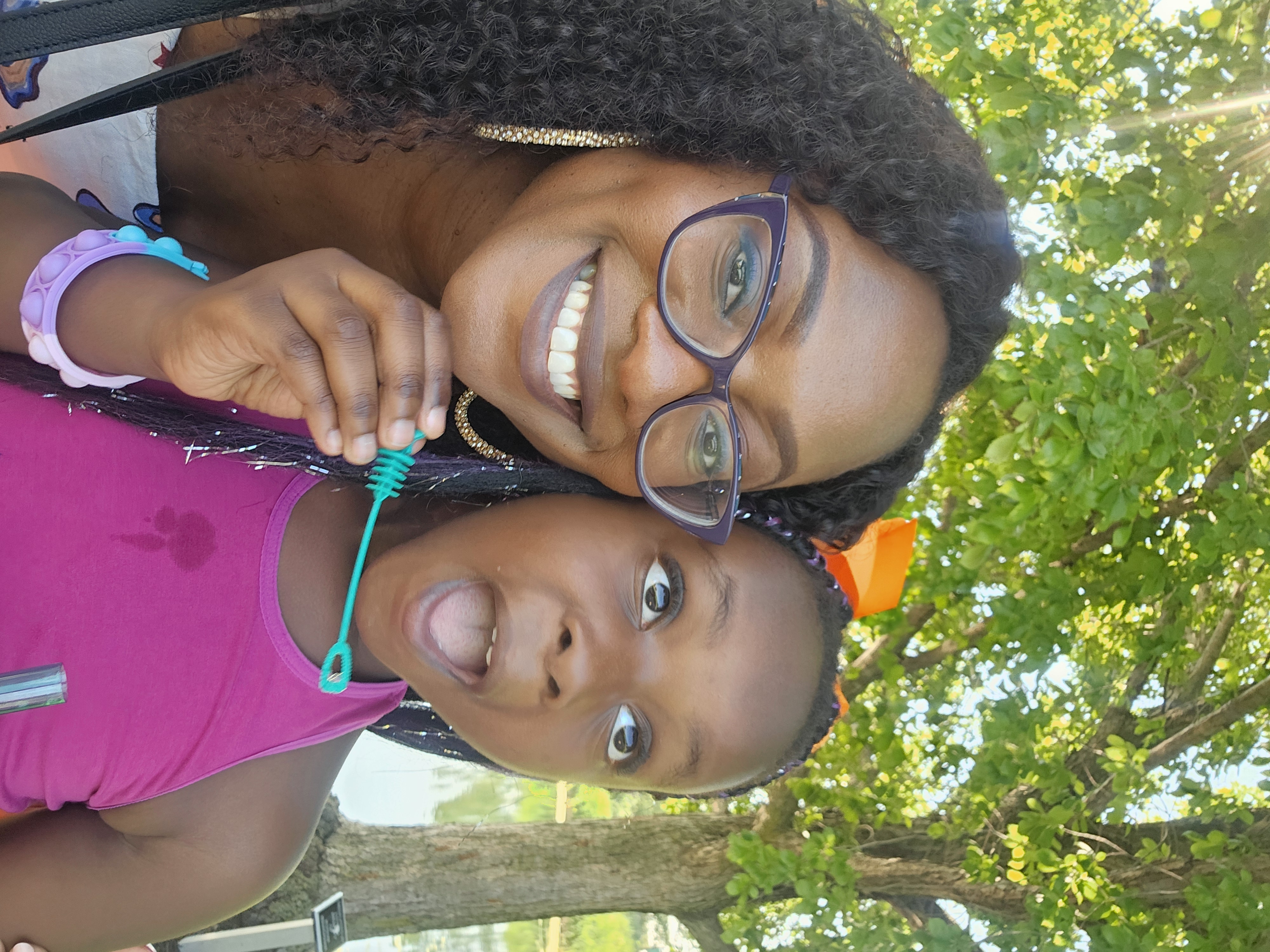 Smiling woman and young girl take a selfie under leafy trees by water; the girl holds a bubble wand and looks surprised.