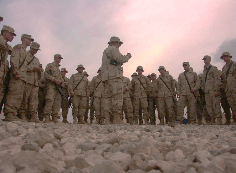 Soldiers in desert camouflage stand on rocky ground as one briefs the group under a pink sunset sky.