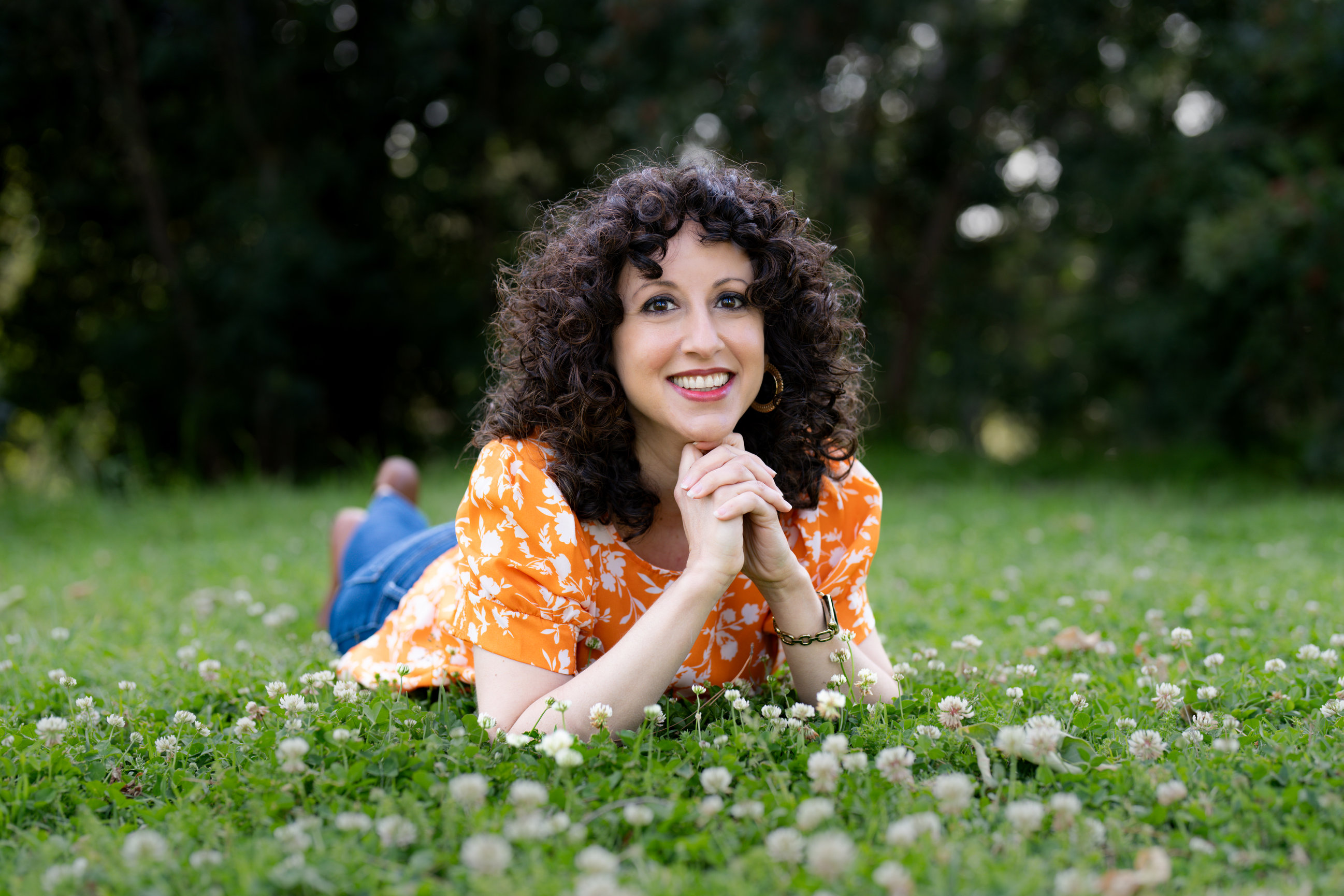 Smiling woman Olivia Abousaid with curly hair lies in a clover-filled park, wearing an orange floral top and jeans.