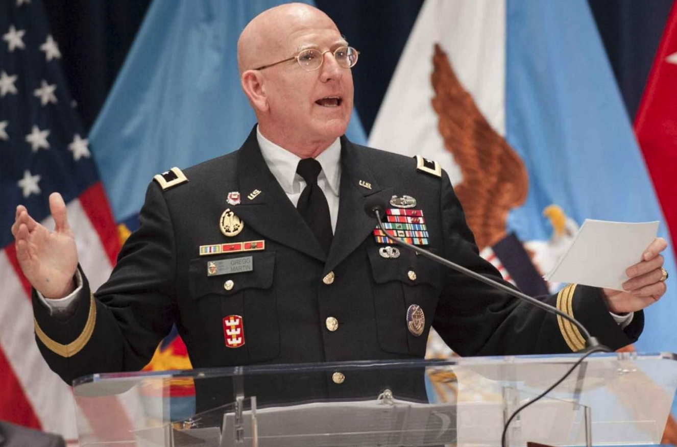 U.S. Army officer in dress uniform speaks at a podium, holding papers, with flags behind him and name tag Gregg Martin.