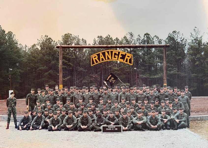 Large group of soldiers in green uniforms pose under a RANGER sign in a pine forest, with a class plaque and flag.