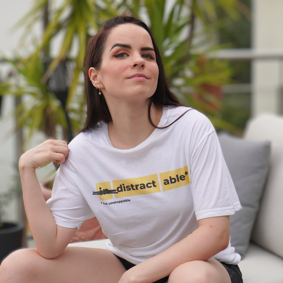 Young woman sitting outdoors in a white T-shirt reading distract able, with potted plants blurred behind her and a calm expression