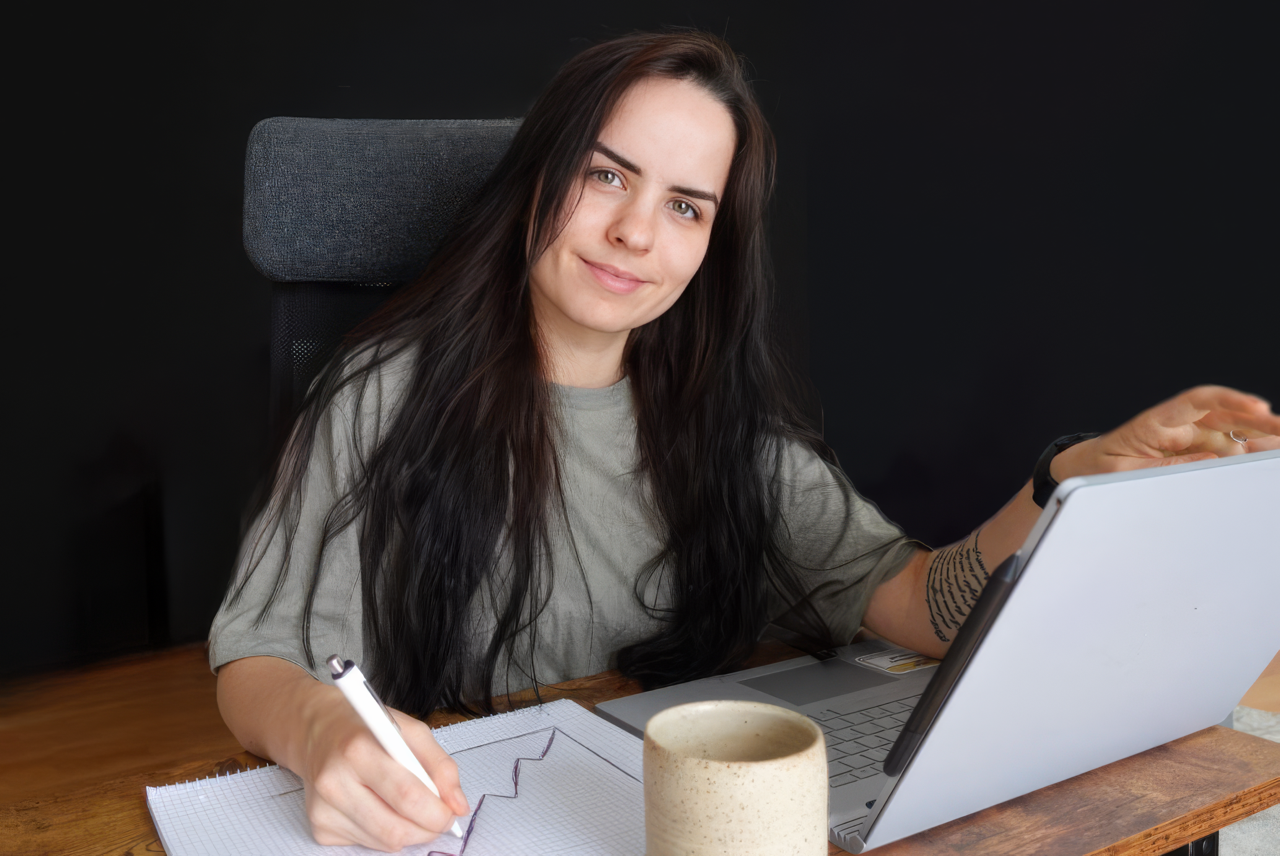 Woman at a desk smiles while writing on graph paper beside an open laptop and mug in a dark home office.