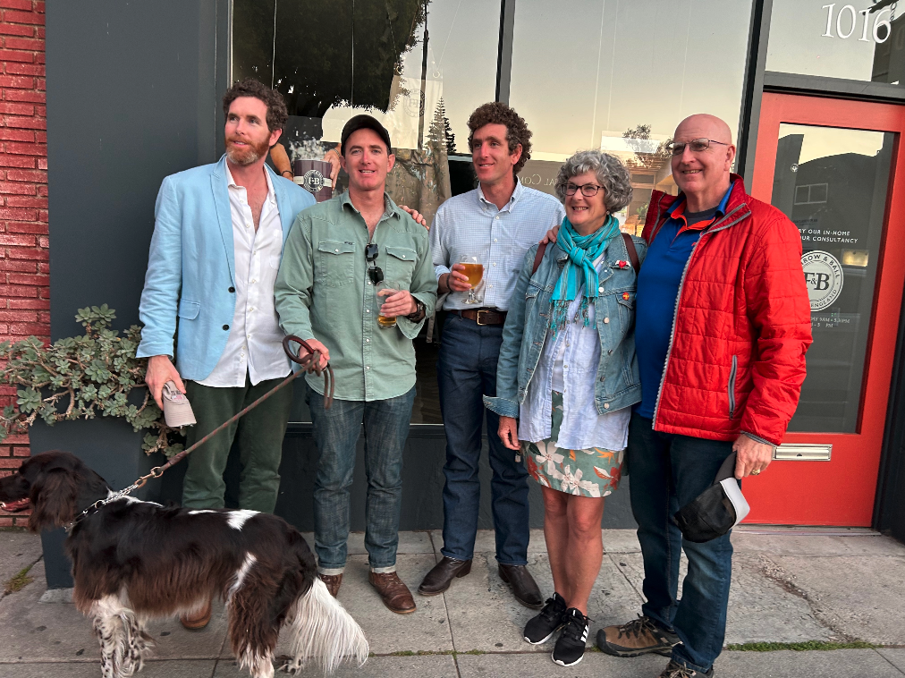 Five smiling adults stand outside a storefront with a dog on a leash; red door marked 1016 and sunset reflections in the window.