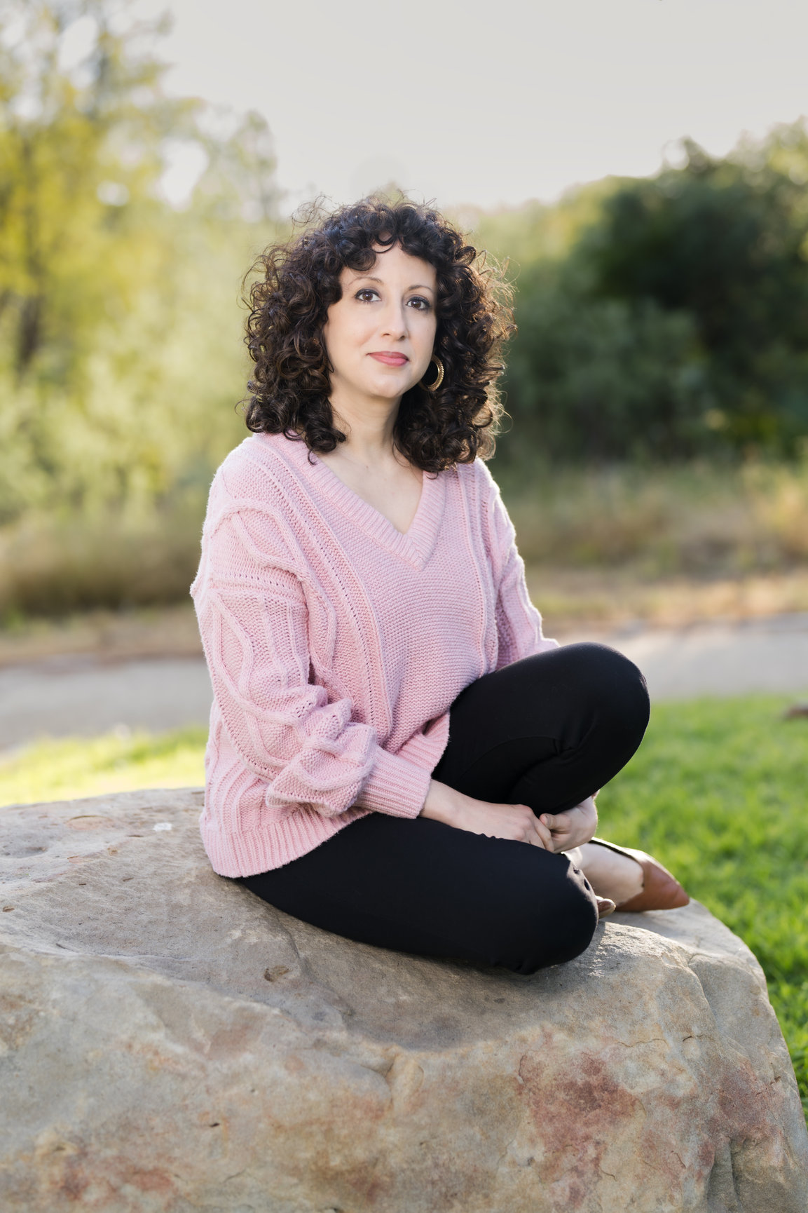 Woman with curly hair Olivia Abousaid in a pink sweater sits cross-legged on a large rock in a sunny park, looking calmly at the camera.