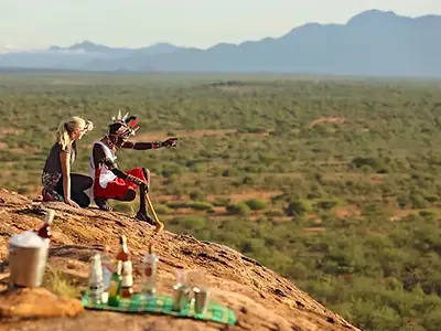 A Samburu warrior in traditional attire points across the vast landscape of northern Kenya, guiding a female guest seated beside him on a rocky outcrop. A picnic with drinks sits nearby, with the green valleys and distant mountains of the Samburu region stretching into the horizon—an authentic cultural experience on an African safari.