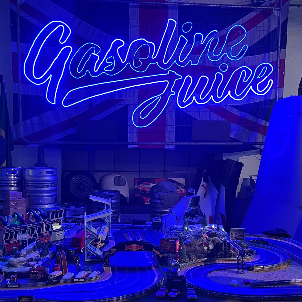 A Scalextric racing setup illuminated by deep blue atmospheric lighting, featuring a large 'Gasoline Juice' neon sign mounted