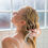 Thumbnail: A blonde woman rinses Tussah Silk & Aloe Vera Shampoo from her hair.  Sunlight enters from a blurred-glass window.