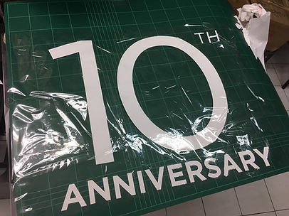 ตัวอักษรสติกเกอร์สีขาวคำว่า "10TH ANNIVERSARY" วางอยู่บนแผ่นตัดสีเขียวในร้านตัดสติกเกอร์ พร้อมแผ่นฟิล์มใสที่คลุมอยู่บางส่วน ข้างโต๊ะมีเศษกระดาษขาวและบรรยากาศของพื้นที่ทำงานผลิตสติกเกอร์