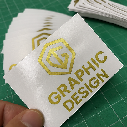 สติ๊กเกอร์พิมพ์สีทองโลโก้และข้อความ 'GRAPHIC DESIGN' บนพื้นขาว เนื้อสติ๊กเกอร์เงา ถูกจัดเรียงซ้อนกันบนพื้นเขียว