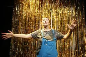 Fran Bushe in overalls, arms wide, in front of strands of curtain in her show Ad Libido