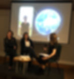 Two authors sitting with the woman interviewing them with a picture of the third author on a screen behind them