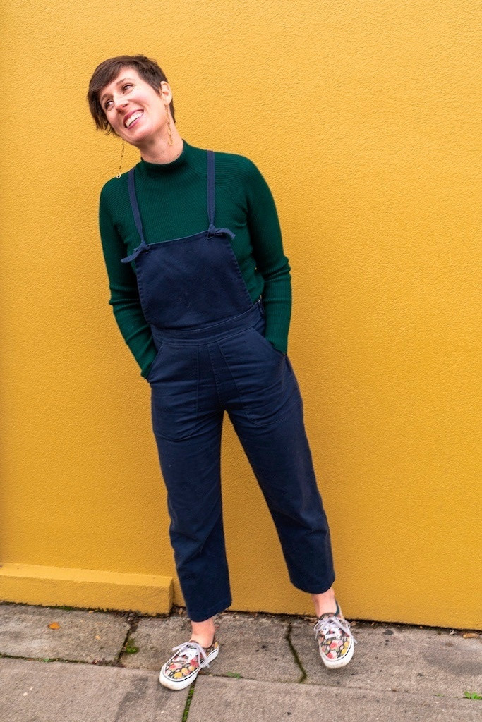 Rebecca Koon, with long earrings and hands in her overalls, smiles and leans to the left