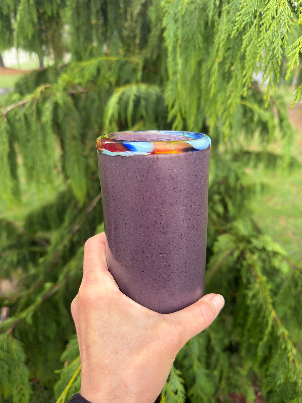 Hand holding a purple smoothie in a cup with a colorful rim against a backdrop of lush green tree foliage.