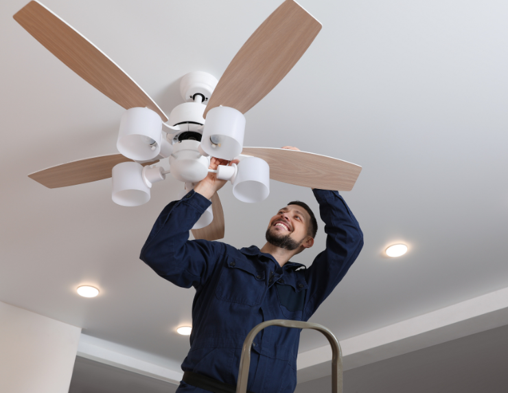 Man in blue overalls on ladder installs a ceiling fan with wooden blades and white fixtures. Bright room with recessed lighting.