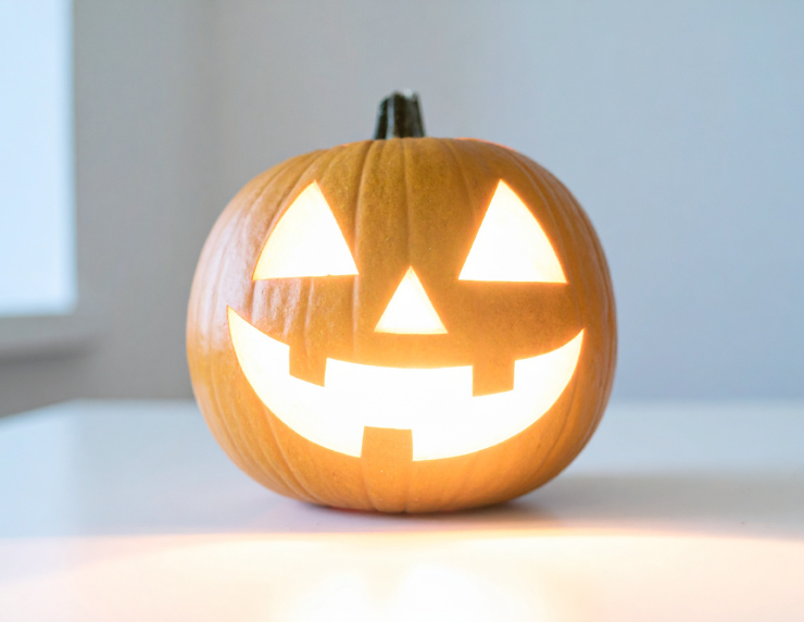 A glowing jack-o'-lantern with a smiling face sits on a white table against a pale blue wall, creating a festive Halloween mood.