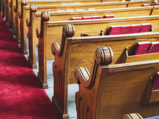 Rows of wooden church pews with red hymnals, seen beside a red carpet aisle in a quiet sanctuary