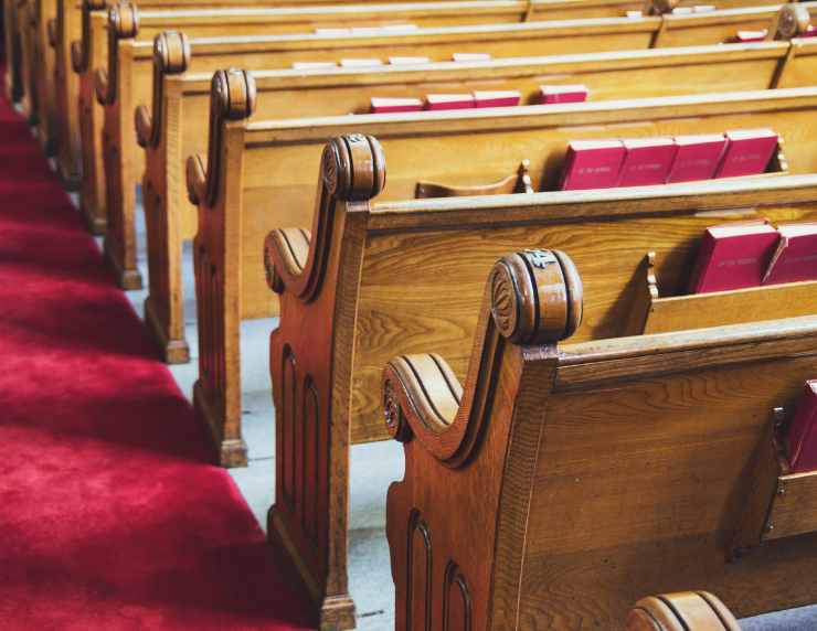 Rows of wooden church pews with red hymnals, seen beside a red carpet aisle in a quiet sanctuary