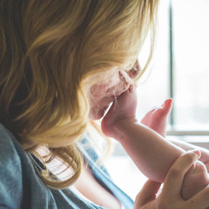 Close-up of a mother kissing and holding her baby’s foot by a bright window, tender and intimate, with soft blond hair visible