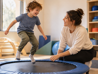 Smiling boy jumps on a blue mini trampoline while a woman kneels nearby, laughing in a bright playroom with exercise gear.