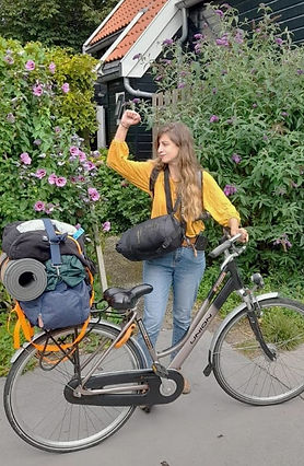 A woman wearing a yellow blouse and jeans stands next to a fully loaded bicycle, raising her fist in a triumphant gesture.